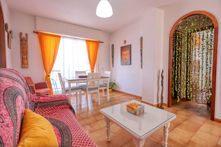 Salón con sofá colorido, mesa central y comedor junto a ventana grande