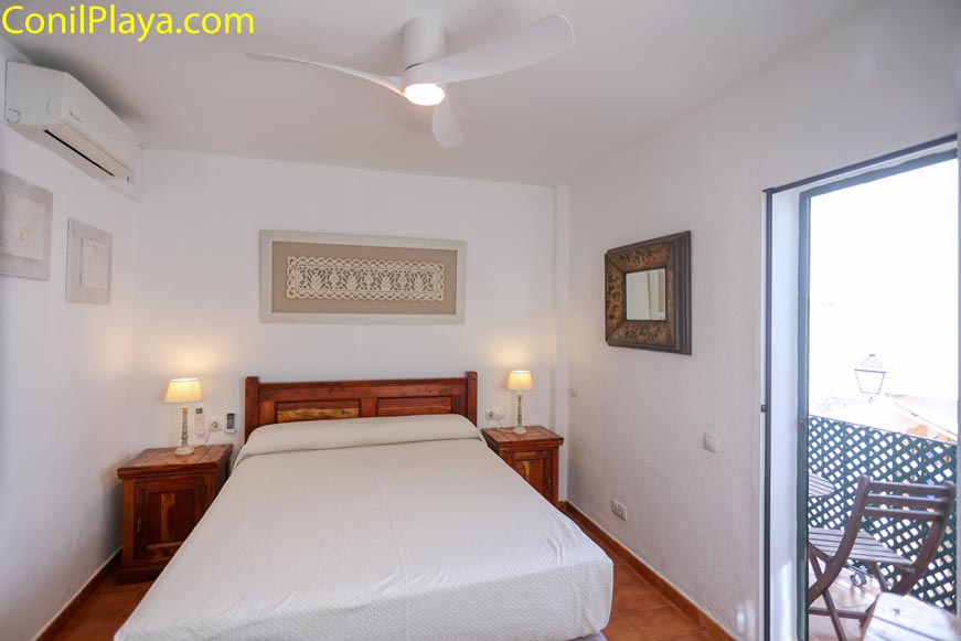 Dormitorio con cama doble, ventilador de techo y aire acondicionado, dos mesitas y acceso a pequeña terraza amueblada