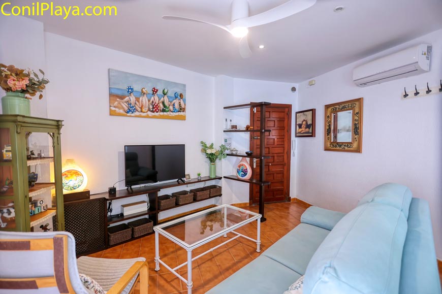 Salón con ventilador de techo y aire acondicionado, con sofá celeste, televisor y mesa de cristal.