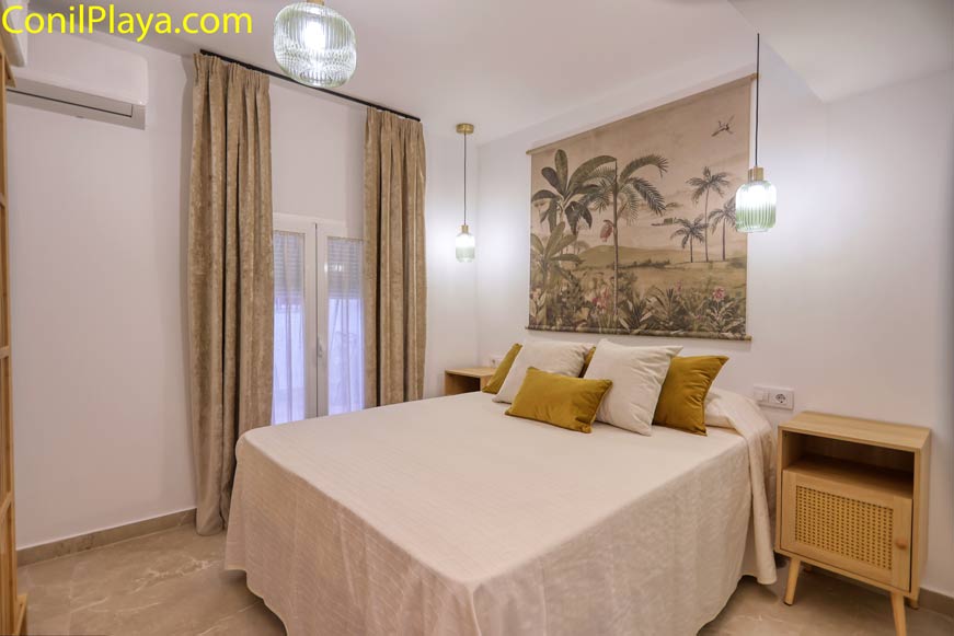 Dormitorio con cama doble, cojines mostaza, mesitas de madera y cuadro grande de palmeras en la pared.