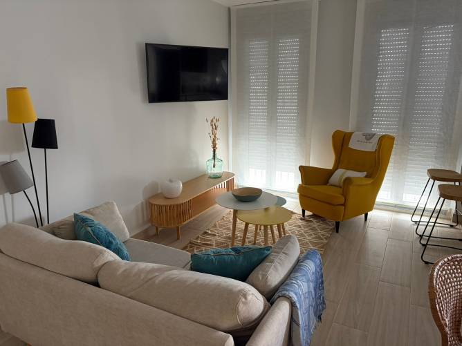 Salón luminoso con sillón orejero amarillo, TV de pantalla plana y ventanales con estores