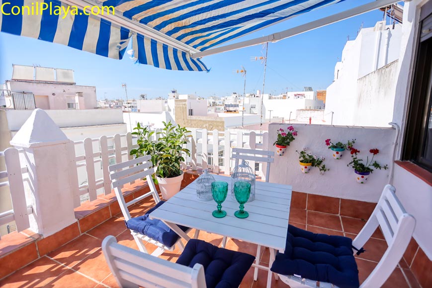 Terraza con mesa blanca y sillas bajo toldo azul rayas y flores en pared