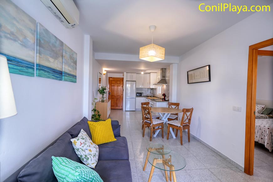 Salón con sofá gris, mesa de comedor y cocina al fondo, entrada a dormitorios