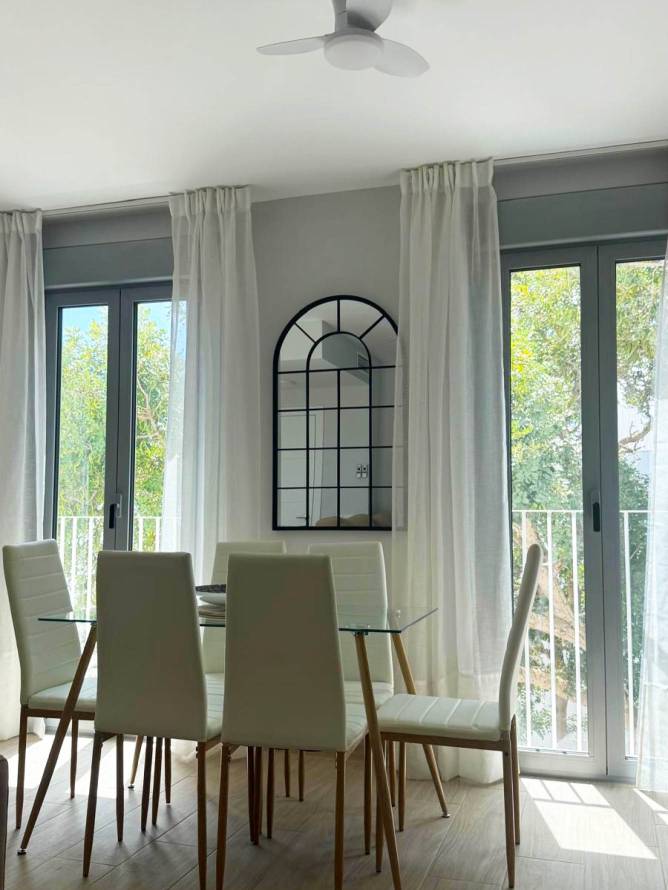 Comedor con mesa de cristal y sillas blancas junto a grandes ventanales y ventilador de techo