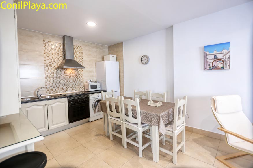 Cocina comedor luminoso con mesa, sillas blancas, electrodomésticos y decoración en la pared.