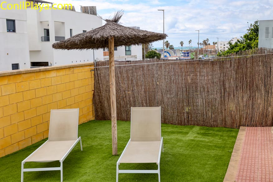 Dos tumbonas y sombrilla de brezo en jardín privado con césped artificial y vallado.