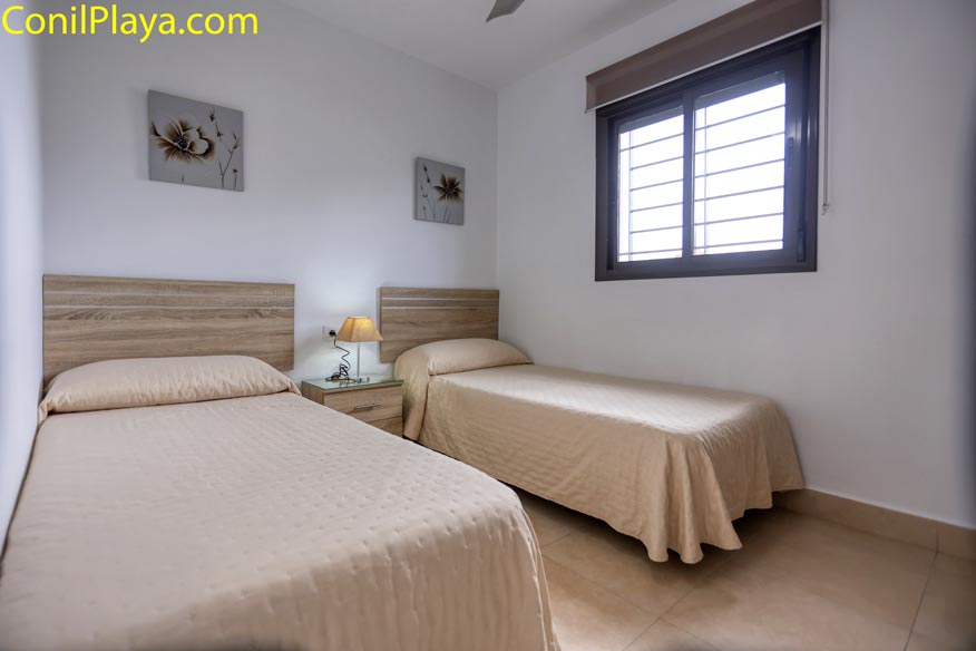 Habitación luminosa con dos camas individuales, ventana, cuadros decorativos y lámpara de noche.