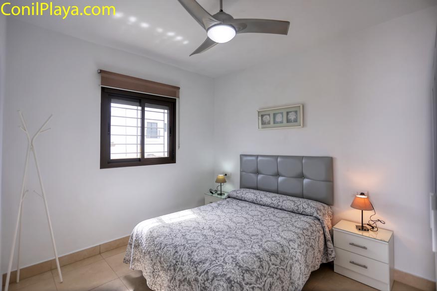 Dormitorio luminoso con cama doble, cabecero gris, ventilador de techo y dos mesitas con lámpara.