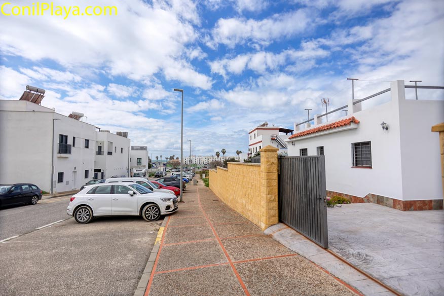Calle residencial con casas blancas, acera amplia y coches estacionados bajo cielo parcialmente nublado.