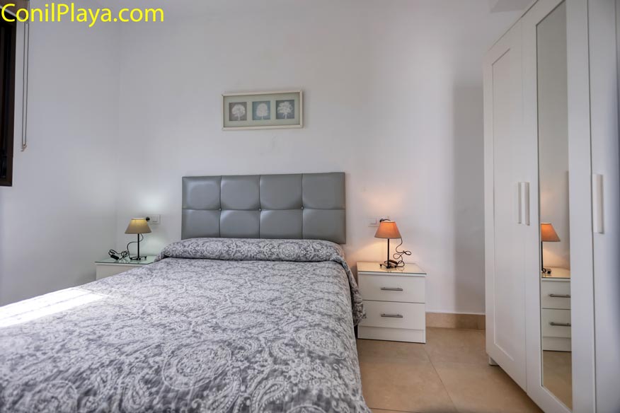Dormitorio con cama doble, cabecero gris, dos mesitas y armario blanco con espejo.