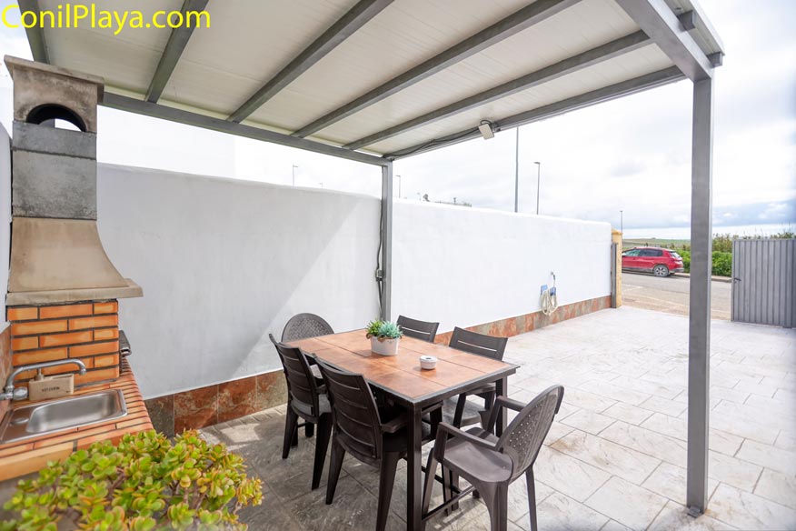 Terraza con mesa, sillas, barbacoa de obra y fregadero bajo techo metálico.
