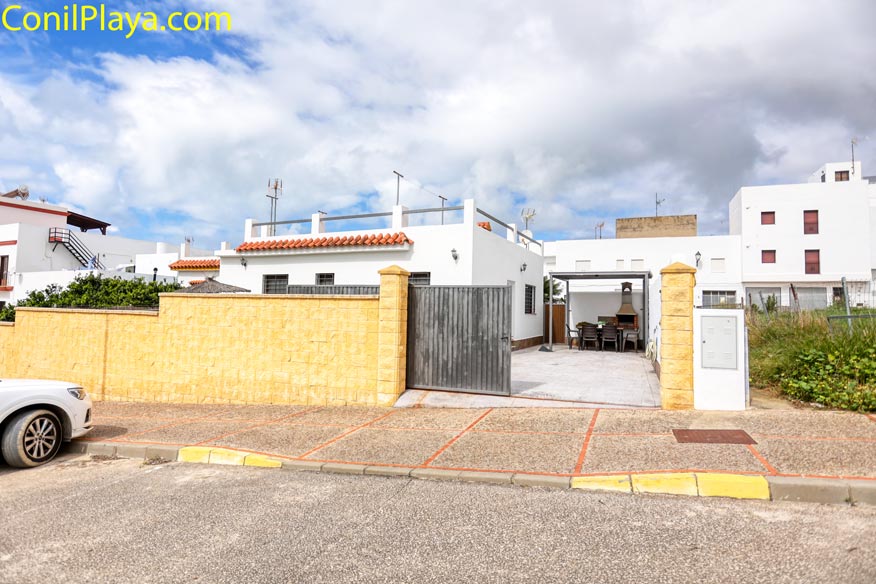 Casa blanca con patio, mesa y sillas exteriores, muro amarillo y portón metálico, cielo nublado.