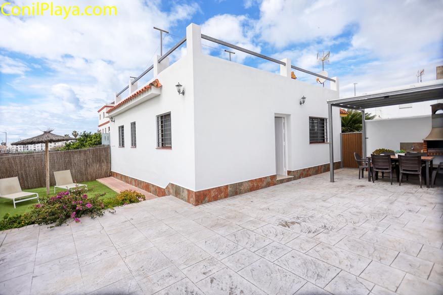 Patio trasero de casa blanca con zona de relax, césped, tumbonas y barbacoa cubierta.