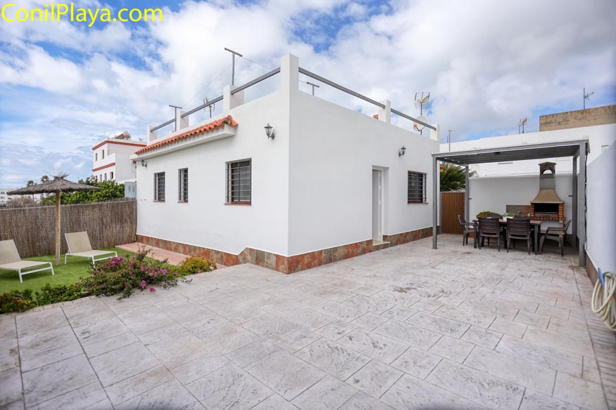 Casa blanca con patio, barbacoa cubierta, mesa exterior y dos tumbonas sobre césped artificial.