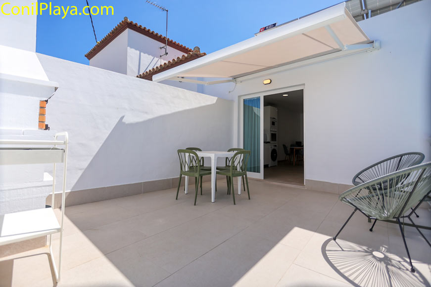 Terraza amplia con mesa y cuatro sillas bajo toldo, junto a pared blanca y acceso a interior de vivienda .