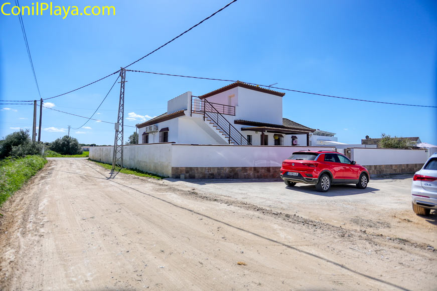 Zona de aparcamiento exterior con dos coches, junto a un edificio blanco y camino de tierra bajo cielo despejado