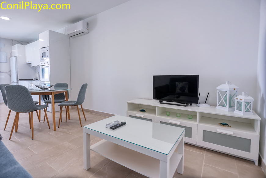 Salón moderno con televisor, mesa de centro de cristal, sillas grises y mueble blanco con decoración