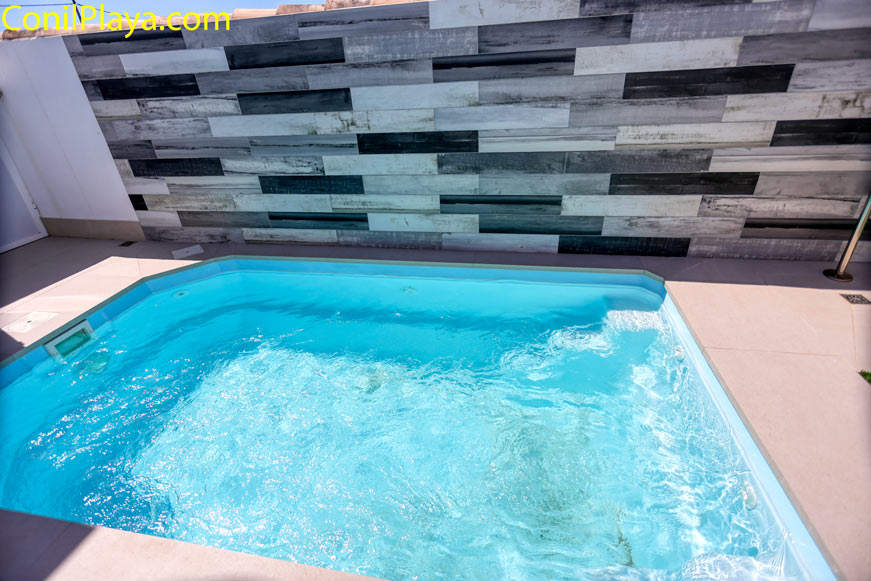 Piscina pequeña y rectangular rodeada de suelo claro junto a una pared decorativa de tonos grises.