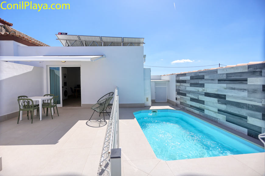 Comedor exterior con mesa blanca y sillas verdes bajo toldo junto a la piscina y la casa en Conil