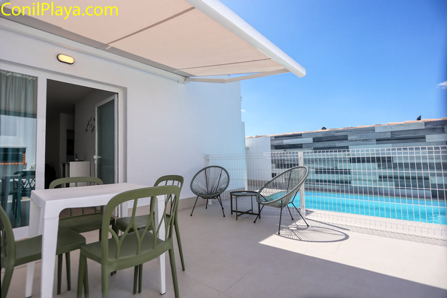 Terraza soleada con mesa, sillas verdes y zona de estar junto a piscina y valla blanca