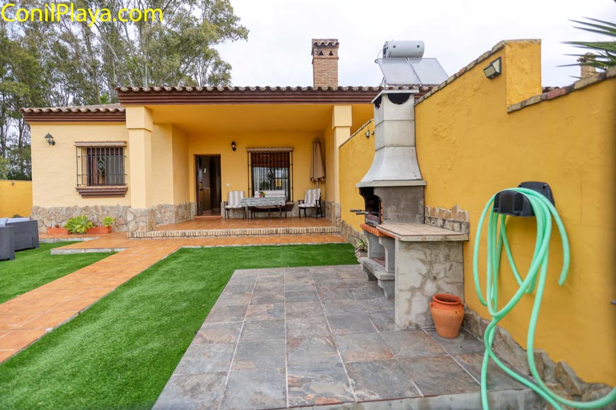 Patio con césped artificial, barbacoa, manguera enrollada y mesa con sillas bajo porche.