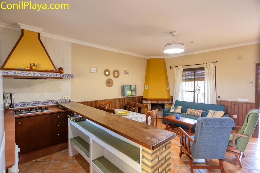 Cocina y salón rústicos con chimenea, barra americana y muebles de madera, ambiente acogedor.