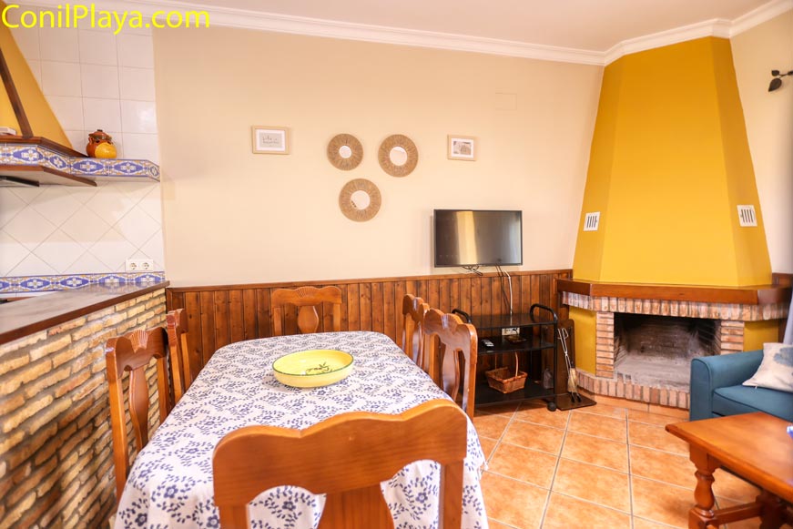 Comedor rústico con chimenea amarilla, mesa de madera y televisor en la pared.