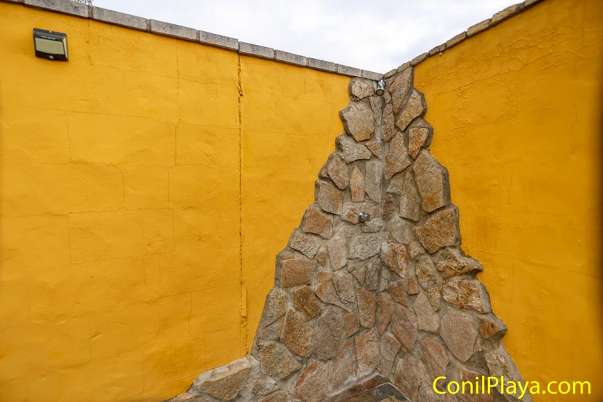 Ducha exterior de piedra en esquina, paredes amarillas y grifo metálico.