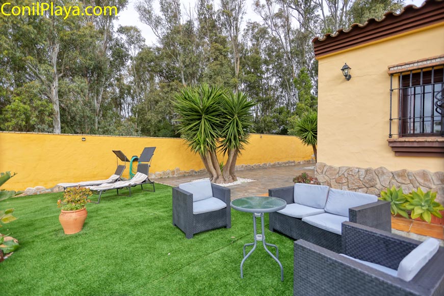 Jardín con césped artificial, muebles de terraza, tumbonas y plantas junto a casa amarilla.