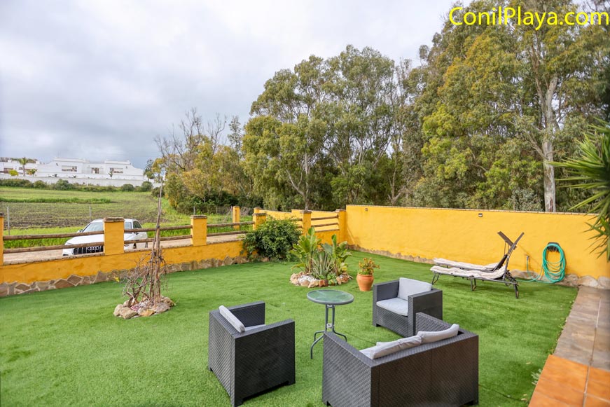 Jardín con césped artificial, muebles exteriores, hamaca y pared amarilla, rodeado de árboles.