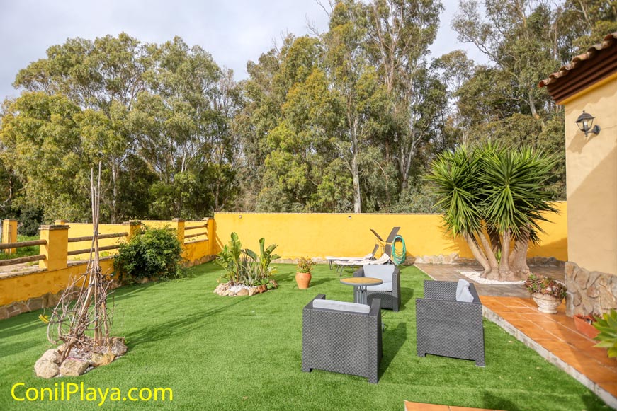 Jardín con césped artificial, muebles exteriores y plantas junto a muro amarillo y árboles.
