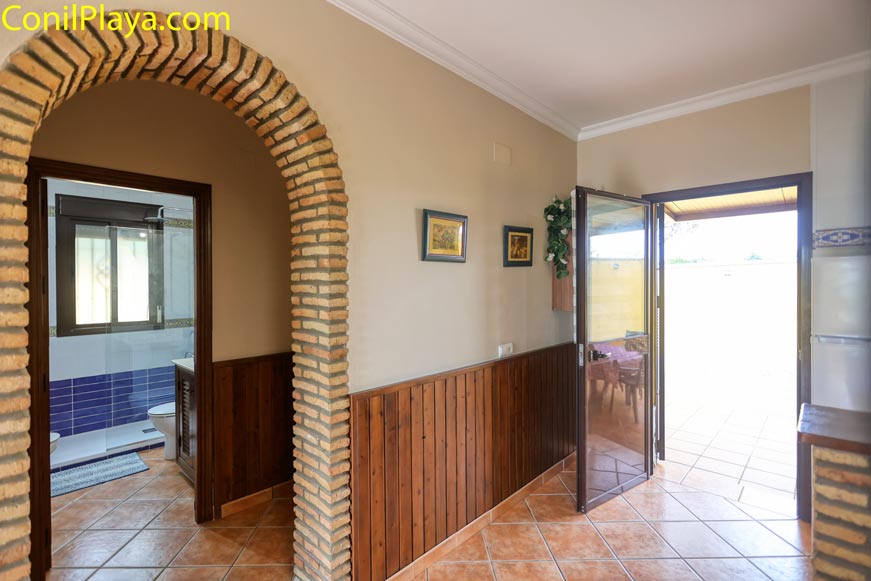 Pasillo con arco de ladrillo, baño al fondo y puerta acristalada hacia terraza exterior.
