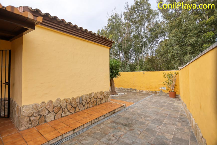 Patio exterior con suelo de baldosas, muro amarillo y plantas en maceta junto a la casa.
