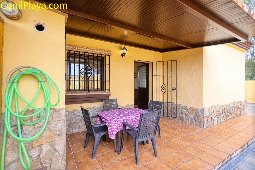 Terraza con mesa y sillas de plástico, manguera enrollada y pared amarilla de piedra.