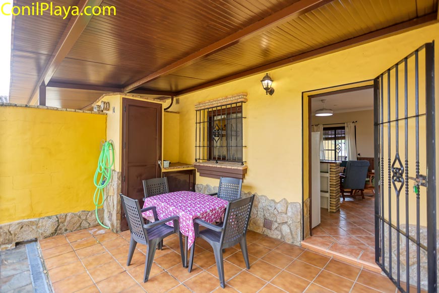 Terraza cubierta con mesa, cuatro sillas de plástico y acceso a sala interior amueblada.