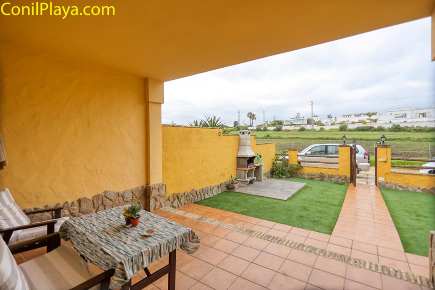 Terraza cubierta con mesa, sillas, barbacoa de obra y jardín con césped al fondo.