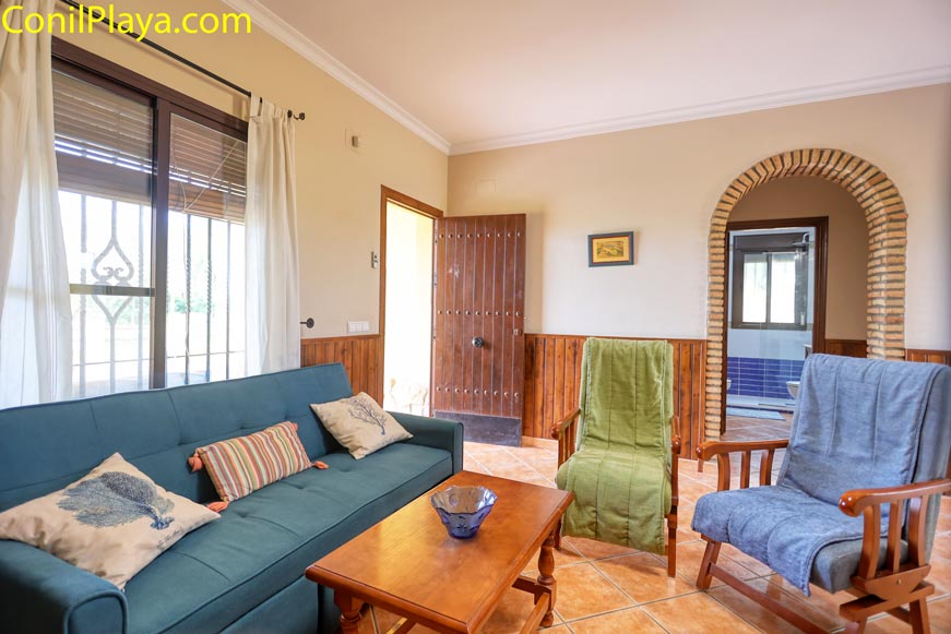 Salón con sofá azul, sillones cubiertos, mesa de madera y puerta de entrada abierta.