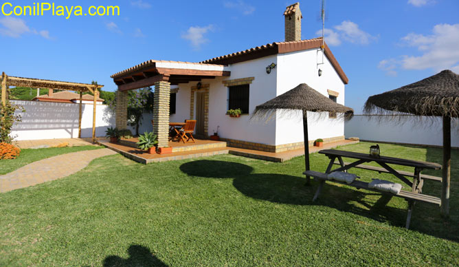 Chalet con porche, jardin con césped amplio y mesa con sombrillas.