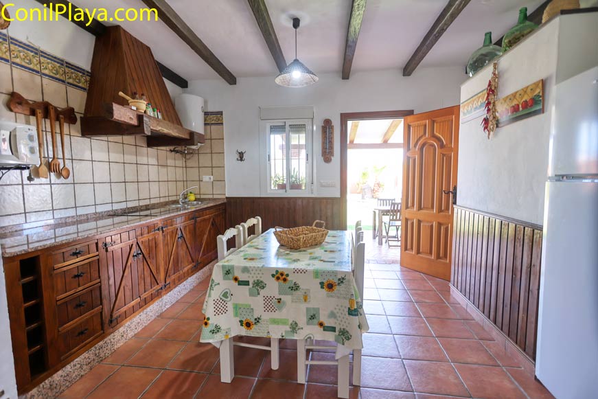 Cocina rústica con mesa central y armarios de madera visible desde el comedor