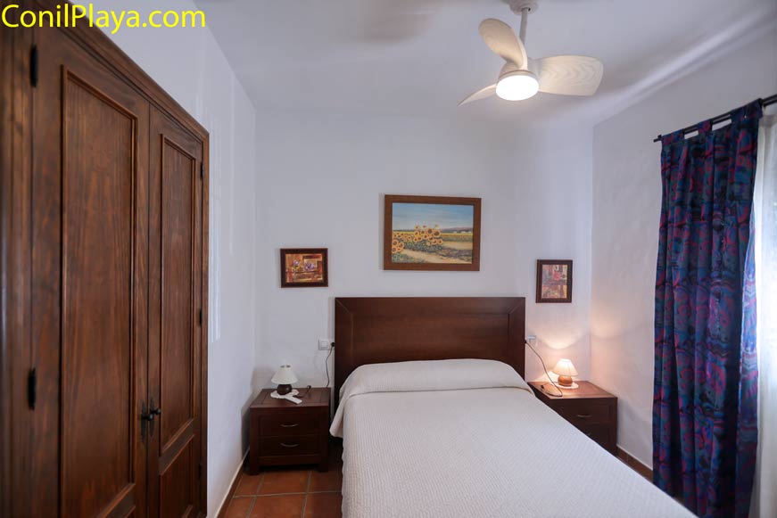 Dormitorio con cama individual, armario de madera, dos mesitas de noche y lámparas, paredes blancas y cortina azul