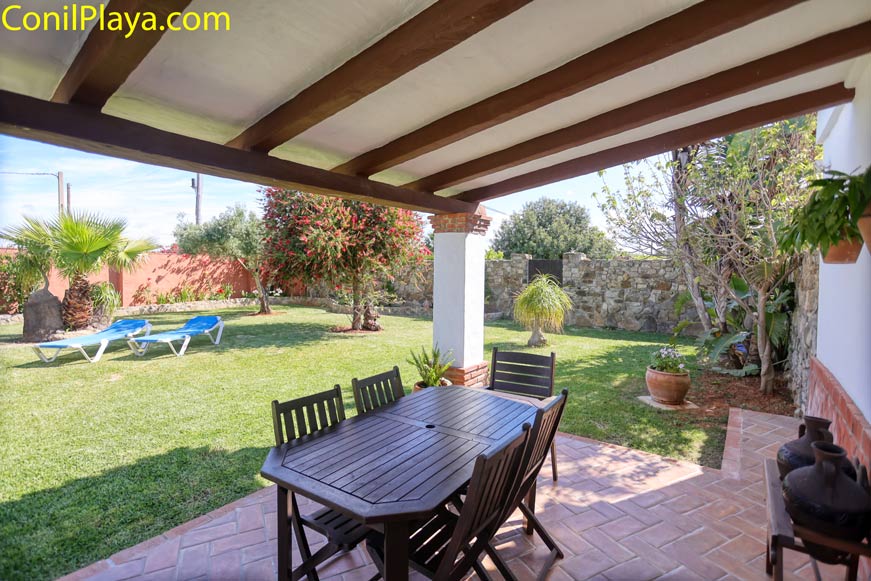 Porche cubierto con mesa y sillas junto a jardín con tumbonas