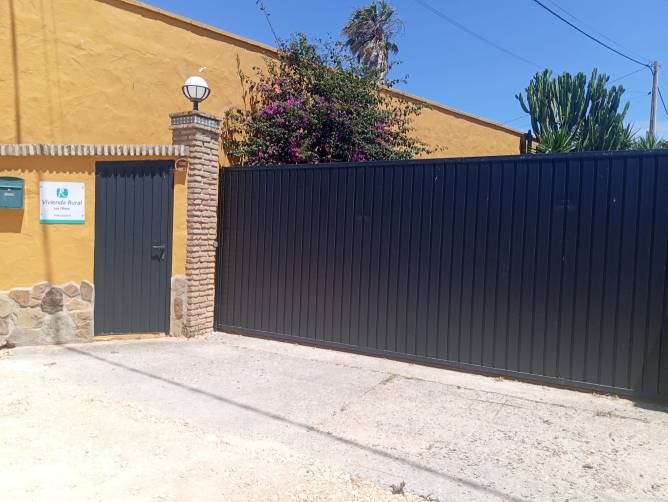 Entrada de aparcamiento con muro amarillo y portón metálico en vivienda rural