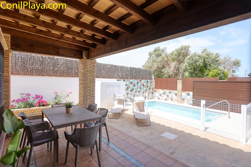 Porche cubierto con mesa y sillas junto a piscina y zona de tumbonas