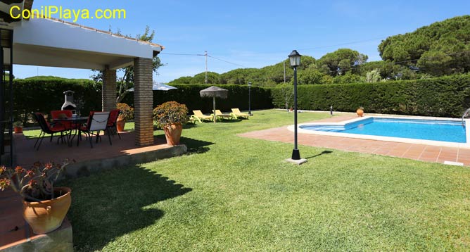 Mesa y sillas del porche y el amplio jardín con la piscina al fondo.