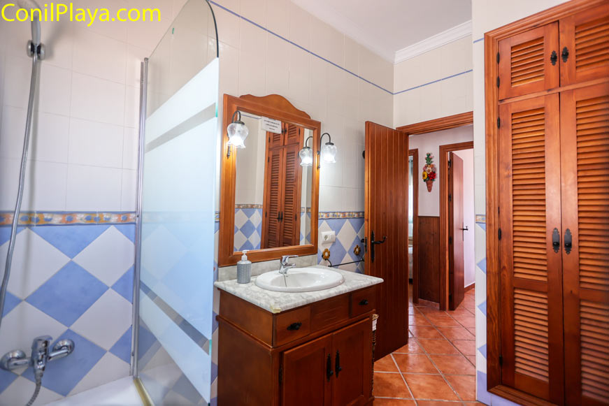 Baño luminoso con ducha, lavabo y armario de madera junto a entrada