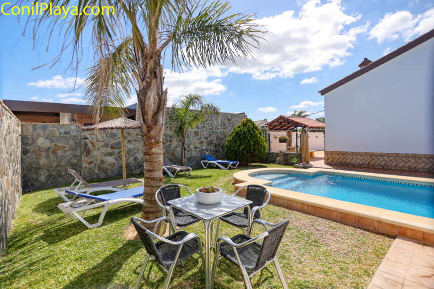 Piscina exterior con jardín, palmera, mesa de cuatro sillas y tumbonas junto a un muro de piedra