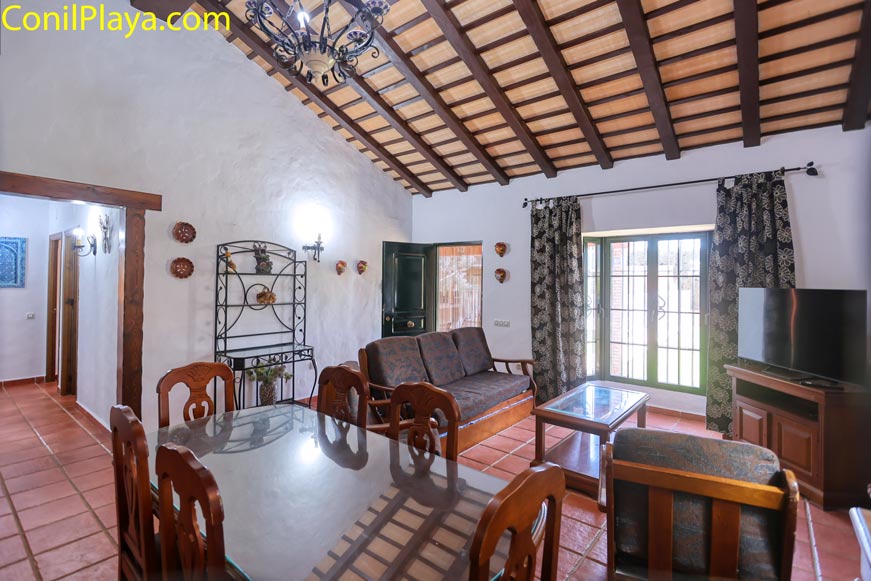 Vista del salón rústico con techo de vigas, sofá, mesa de comedor y televisor