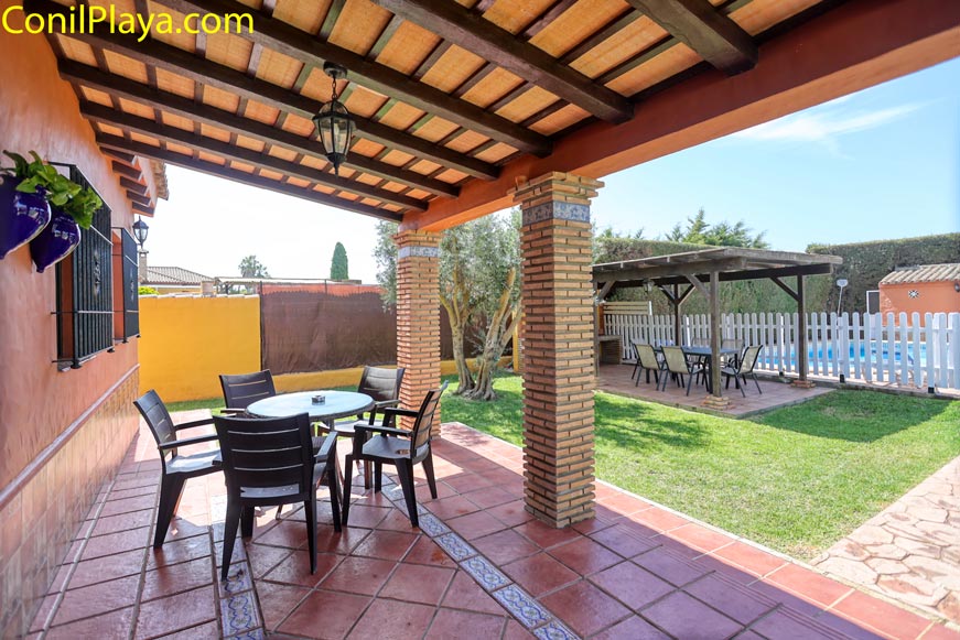 Porche cubierto con mesa junto a jardín y zona de piscina vallada