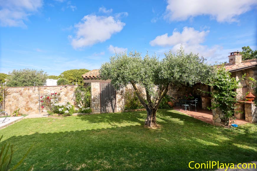 Jardín con césped, un árbol central, setos, flores junto a muro de piedra y porche cubierto con mesa y sillas