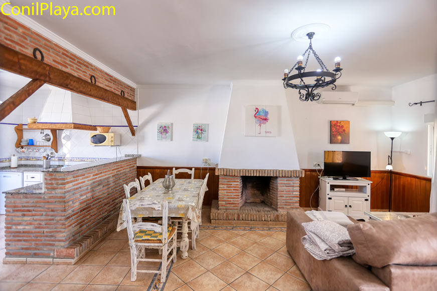 salón con chimenea, sofá, mesa de comedor y cocina integrada de ladrillo